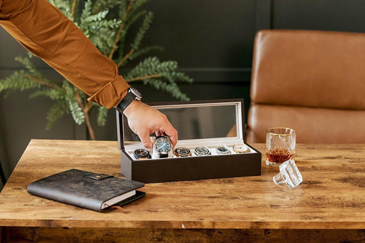 Person selecting a watch from a display case on a wooden table with a glass of whiskey.