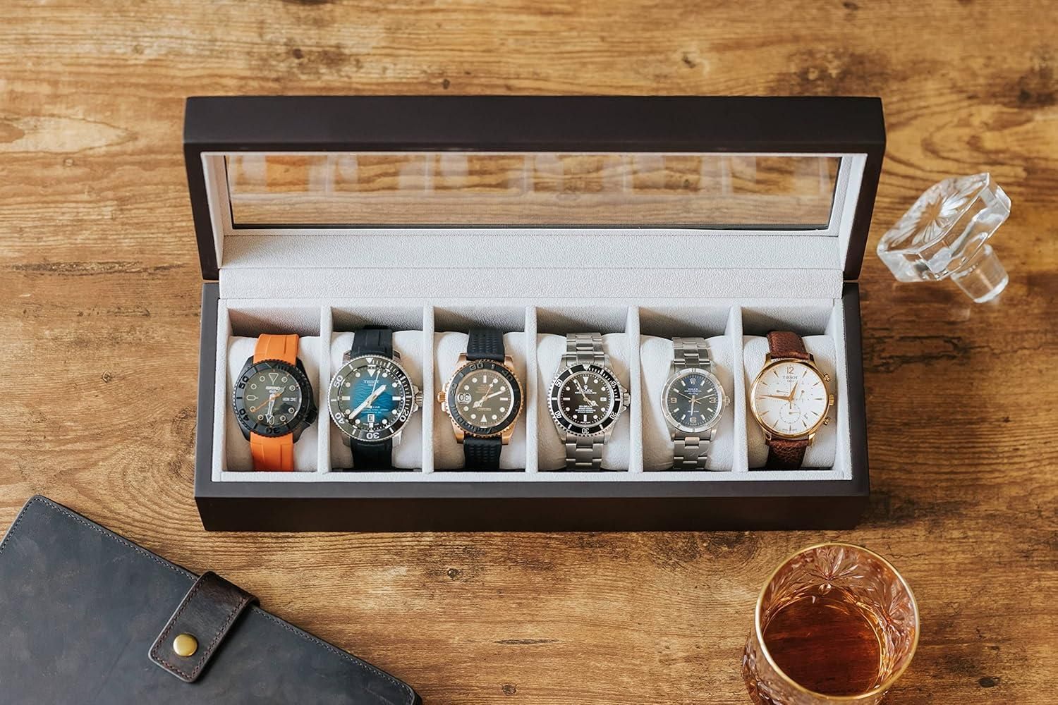 Set of six watches in a display case on a wooden surface with glasses and a leather case.