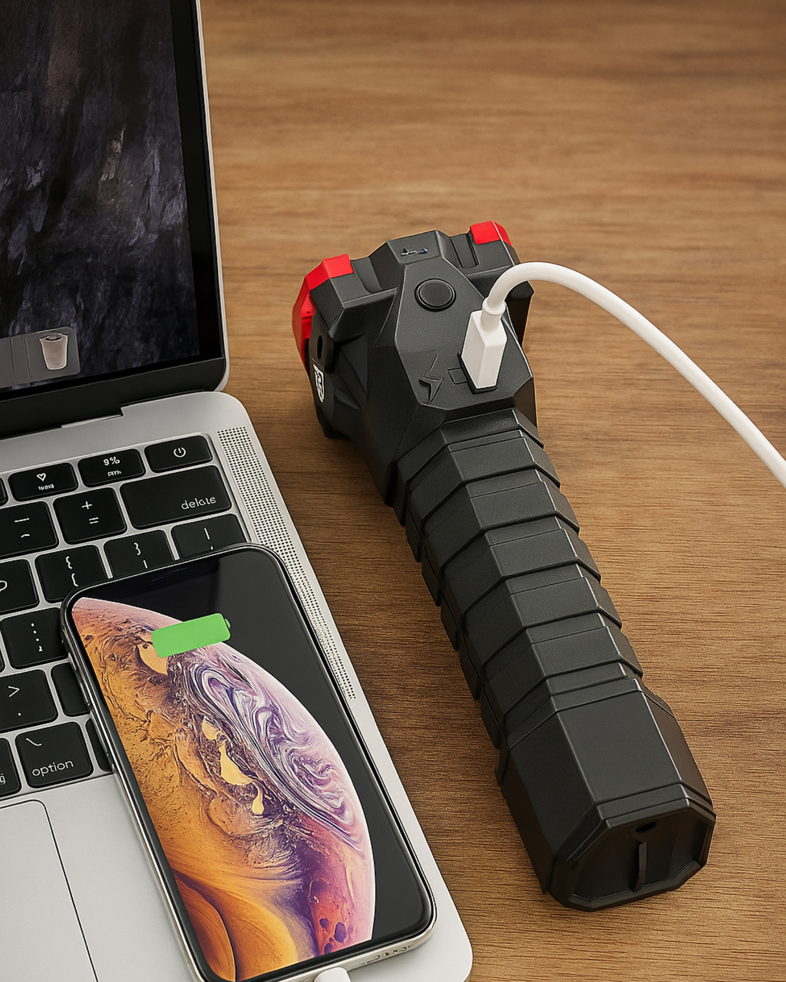 Black power bank with white cable on a wooden surface next to a laptop and smartphone.